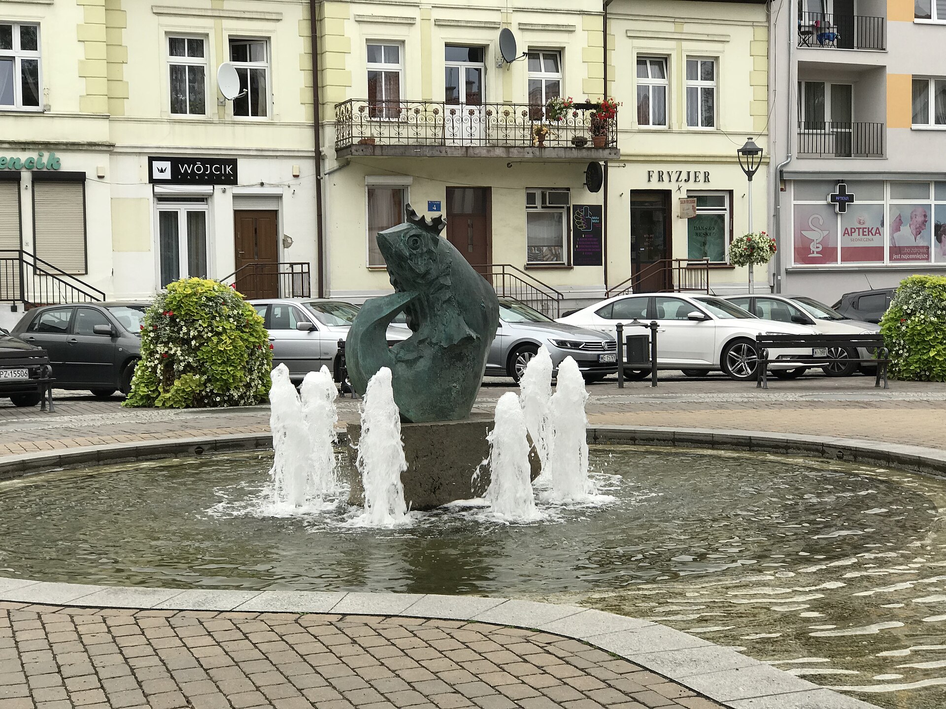 Zielona rzeźba ryby w koronie stojąca w fontannie, w tle kamienice w centrum Mikołajek